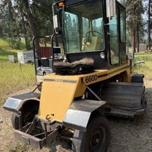 5 2 - Montana Auctions by Kevin Hill Yellow road roller with a large front broom sweeper, parked on dirt beside trees in a rural lot.