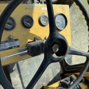 5 8 - Montana Auctions by Kevin Hill Close-up of a yellow industrial machine cab with a large black steering wheel and four round gauges on the dashboard, plus levers nearby.