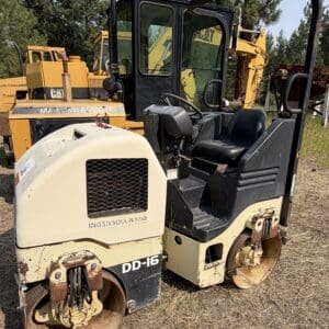 6 1 - Montana Auctions by Kevin Hill Front view of a small double-drum road roller (DD-16) in beige and black, parked on a grassy lot with yellow construction equipment in the background.