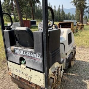 6 1 - Montana Auctions by Kevin Hill Back view of a small ride-on asphalt roller in a grassy field, with trees and construction gear in the background; the drum shows rust.