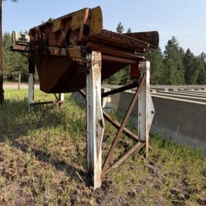 7 1 - Montana Auctions by Kevin Hill Old, rusted industrial conveyor/machine on a metal frame outdoors with grass and trees in the background.