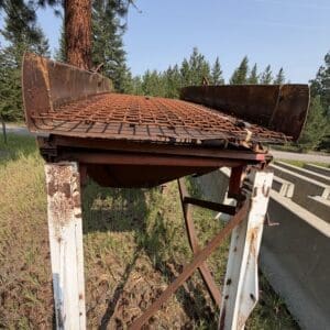 7 1 - Montana Auctions by Kevin Hill Old rusted metal platform on white legs beside a road, in a pine forest setting.