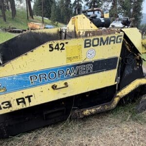 8 1 - Montana Auctions by Kevin Hill Yellow Bomag PROPAVER asphalt paver lying on a grassy field, weathered with rust and dirt, model numbers 542 and 813 RT visible on the side.
