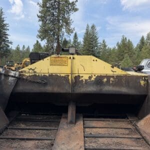 8 3 - Montana Auctions by Kevin Hill Front view of a weathered yellow industrial conveyor with rust, outdoors among pine trees under a blue sky.