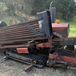 9 3 - Montana Auctions by Kevin Hill Old red agricultural/industrial machine with tracks, sitting on dry grass in a field, showing metal rollers and a conveyor-like frame.
