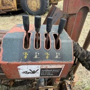 9 3 - Montana Auctions by Kevin Hill Five black levers on a rusty red control console with a yellow warning label about a turning shaft; a large off‑road tire is in the background.