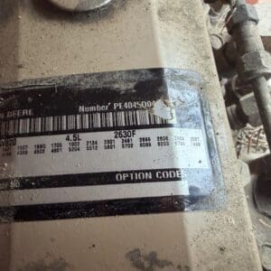 9 5 - Montana Auctions by Kevin Hill Close-up of a dusty machine label showing model and option codes on a John Deere device with a barcode block beneath the dirt.