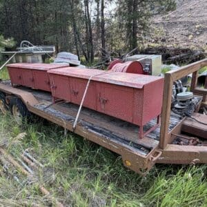 IMG_8526 - Montana Auctions by Kevin Hill Rusty flatbed trailer in a grassy clearing with red diamond-plate tool boxes and scattered equipment.