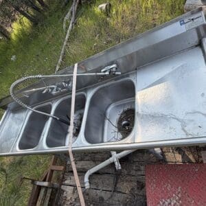 IMG_8528 - Montana Auctions by Kevin Hill Triple-compartment stainless steel utility sink on a trailer, with a sprayer; one basin contains a bird's nest and twigs (outdoors).