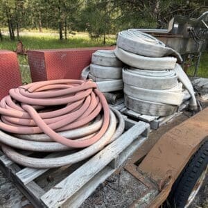 IMG_8529 - Montana Auctions by Kevin Hill Coiled pink and gray hoses on a weathered wooden pallet on a rusted utility trailer in a grassy, wooded area outside.
