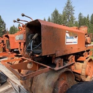 IMG_8532 - Montana Auctions by Kevin Hill Rusty orange Rammax Trampler tracked construction machine on a trailer, dirty with mud, in a grassy, forested area outside.
