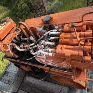 IMG_8534 - Montana Auctions by Kevin Hill Close-up of an orange hydraulic pump system with tangled hoses and worn metal, mounted on a wooden platform outdoors.
