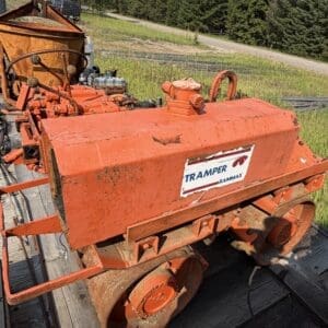 IMG_8540 - Montana Auctions by Kevin Hill Orange tracked roller/compactor on a flatbed trailer, rusted and dusty, with a 'TRAMPER RAMMAX' label in a grassy yard.
