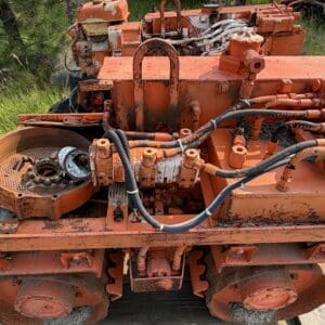 IMG_8542 - Montana Auctions by Kevin Hill Rusty orange industrial machine with hydraulic hoses and exposed gears, sitting on a flatbed outdoors amidst tall grass (abandoned).