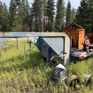 IMG_8543 - Montana Auctions by Kevin Hill Rusty flatbed trailer with orange industrial equipment parked in tall grass beside a forest.