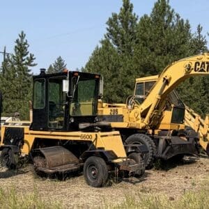 IMG_8554 - Montana Auctions by Kevin Hill Yellow construction excavator and other heavy equipment parked in a sunlit grassy lot with trees in the background.
