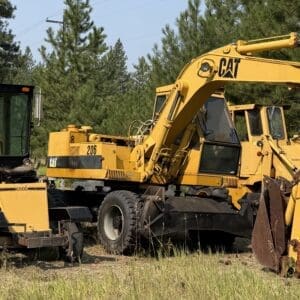 IMG_8555 - Montana Auctions by Kevin Hill Yellow CAT excavator and a road roller sit idle in a grassy field with trees in the background, an outdoor equipment yard scene.