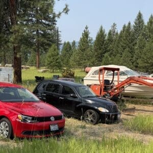 IMG_8687 - Montana Auctions by Kevin Hill Red sedan and black SUV parked on a grassy lot with a boat on a trailer and an orange excavator in the background in a rural salvage yard.