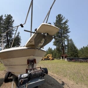 IMG_8724 - Montana Auctions by Kevin Hill White motorboat on a trailer in a grassy open area, with a winch and anchor gear in the foreground and trees in the background.