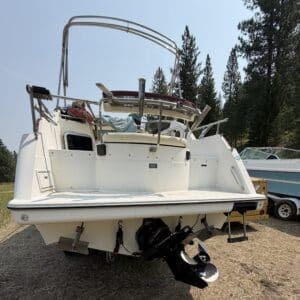 IMG_8725 - Montana Auctions by Kevin Hill Back view of a white motorboat on a trailer, showing the stern with two large outboard motors hanging below and a covered cockpit area in an outdoor boat yard.
