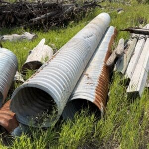 IMG_8774 - Montana Auctions by Kevin Hill Pile of corrugated metal pipes and weathered wooden planks lying on tall grass in an outdoor yard area.