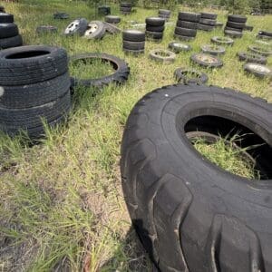 IMG_8835 - Montana Auctions by Kevin Hill Sunlit field filled with used tires of different sizes stacked and scattered across tall grass, with a large tire in the foreground open-ended.