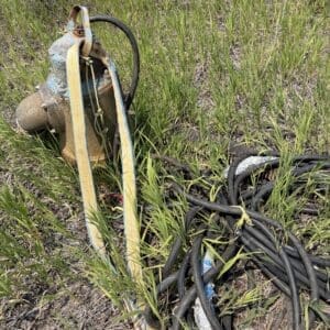 IMG_8838 - Montana Auctions by Kevin Hill Old rusty irrigation pump with yellow straps and a thick black hose, resting in a field of tall green grass.