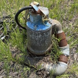 IMG_8839 - Montana Auctions by Kevin Hill Old, rusted submersible water pump in a grassy field with tangled hoses nearby.