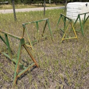 IMG_8845 - Montana Auctions by Kevin Hill Green and yellow metal sawhorses arranged as a makeshift barrier in a grassy field near a fence and a white plastic water tank in the background.