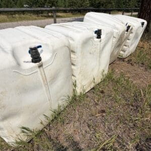 IMG_8847 - Montana Auctions by Kevin Hill Row of white plastic water containers with blue taps along a fence in a grassy area