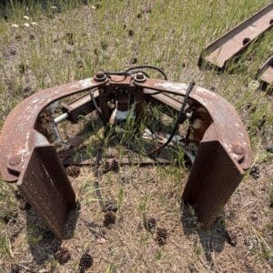 IMG_8849 - Montana Auctions by Kevin Hill Abandoned rusted circular metal machinery frame in a field with pine cones and overgrown grass, with rails stacked in the background.