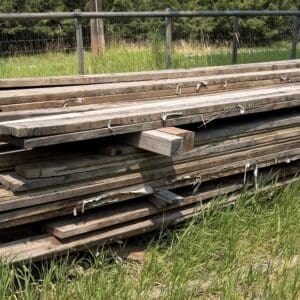 IMG_8852 - Montana Auctions by Kevin Hill Stack of weathered wooden planks outdoors on a grassy field beside a wire fence in the background.