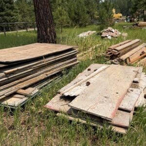 IMG_8853 - Montana Auctions by Kevin Hill Stacks of weathered plywood sheets and lumber piled in a grassy field at a construction site, with trees and distant equipment in the background.