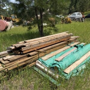 IMG_8855 - Montana Auctions by Kevin Hill Stacks of weathered wooden planks piled on grass with a green tarp nearby; outdoor yard with machinery in the background.