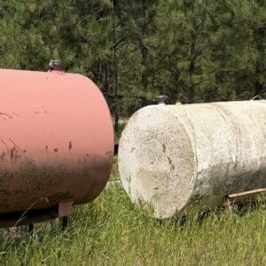 IMG_8856 - Montana Auctions by Kevin Hill Two large cylindrical storage tanks sit on grassy field; left tank is pink and rusted, right tank is white and weathered with trees in the background.
