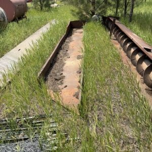 IMG_8861 - Montana Auctions by Kevin Hill Overgrown yard with rusted metal troughs and long, weathered machinery in a sunny, tree-filled field.