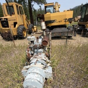 IMG_8865 - Montana Auctions by Kevin Hill Rusty old industrial engine/pump lying in a grassy field, with large yellow construction machines in the background.