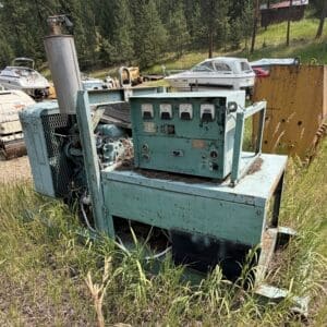Old teal industrial generator in a grassy field, rusted and partially overgrown, with boats and a yellow rusted container in the background.]