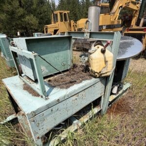 IMG_8869 - Montana Auctions by Kevin Hill Abandoned rusted blue-green industrial machine outdoors with overgrown grass and construction vehicles in the background.