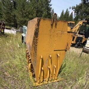 IMG_8875 - Montana Auctions by Kevin Hill Yellow, rusted hydraulic attachment in a grassy salvage yard with other heavy equipment in the background.