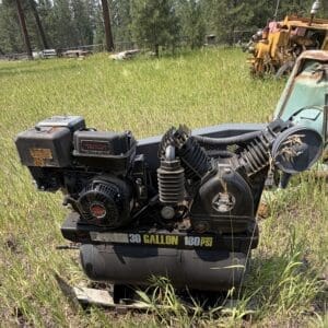 IMG_8882 - Montana Auctions by Kevin Hill Abandoned green and rusted tractor attachment in tall grass, with a yellow hydraulic unit and pine trees in the background.