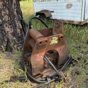 IMG_8883 - Montana Auctions by Kevin Hill Rusted hydraulic attachment on the ground beside a tree, with tangled hoses and a weathered trailer in the background.