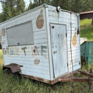 IMG_8884 - Montana Auctions by Kevin Hill Weathered white travel trailer with rust and faded logos in a grassy, tree-filled field in the outdoors.