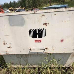 IMG_8888 - Montana Auctions by Kevin Hill White rusted equipment container outdoors with peeling paint and a black geometric logo, grasses around the base, and boats/vehicles in the background