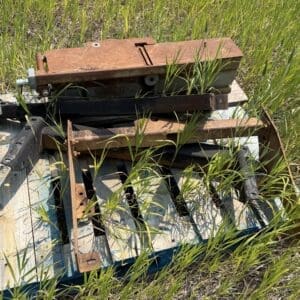 IMG_8911 - Montana Auctions by Kevin Hill Rusty, disused metal machinery piled in a grassy field, with weeds growing through and around the pieces.