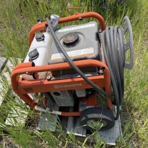 IMG_8912 - Montana Auctions by Kevin Hill Portable orange Husqvarna generator with fuel tank and black hose, sitting on a wooden pallet in tall grass outdoors.