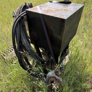 IMG_8914 - Montana Auctions by Kevin Hill Locked rusty metal electrical box with thick cables coiled around it in a grassy field; appears abandoned outdoor equipment.