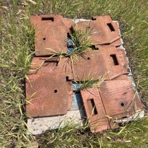 IMG_8925 - Montana Auctions by Kevin Hill Rusty red metal manhole covers stacked on a wooden pallet in a grassy field, with weeds growing between them.