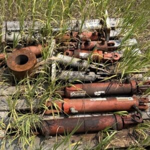IMG_8926 - Montana Auctions by Kevin Hill Rusty hydraulic cylinders and metal fittings piled on a wooden pallet, overgrown with tall grass in a field setting.