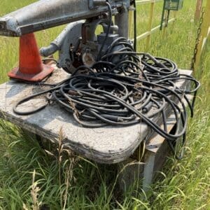 IMG_8930 - Montana Auctions by Kevin Hill Old industrial hydraulic pump on a weathered platform with a tangle of black cables, outdoors in tall grass beside a fence and an orange cone nearby.